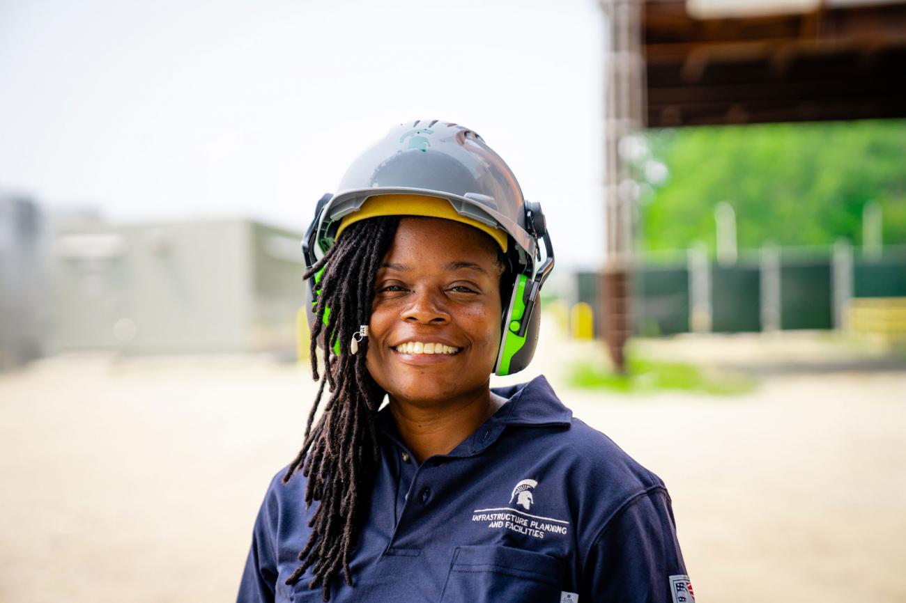 Cher Briggs, a black woman with asymmetrical locs, smiles at the camera in a hard hat and ear protection