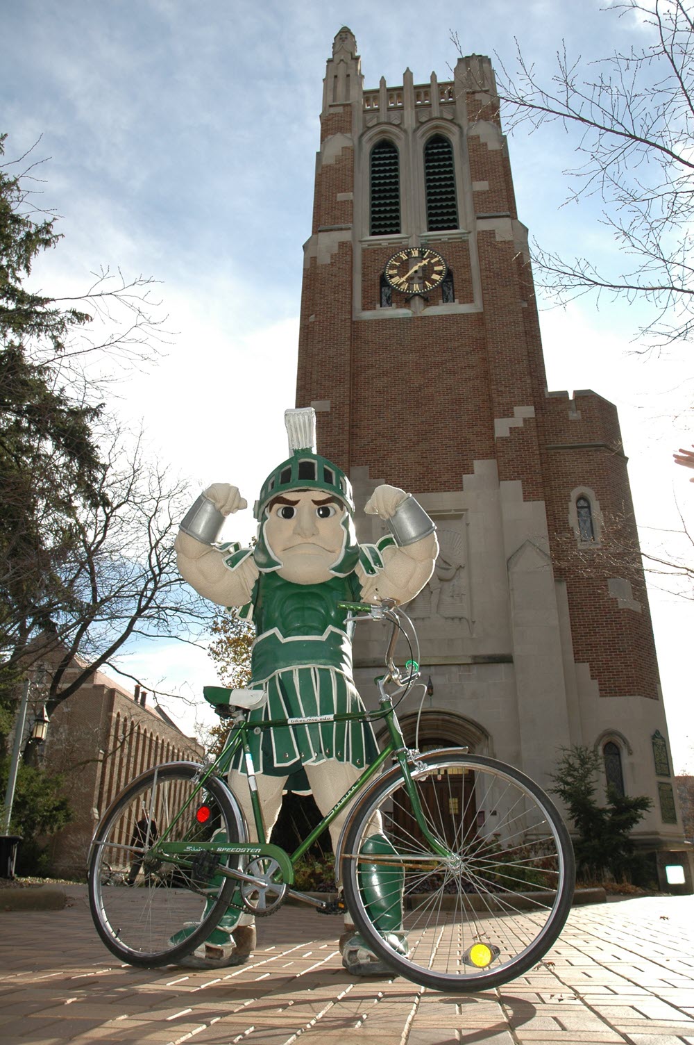 Sparty standing with bicycle in front of Beaumont Tower