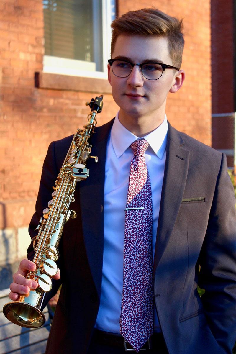 Noah stands wearing a suit and horn-rimmed glasses and holding a brass instrument