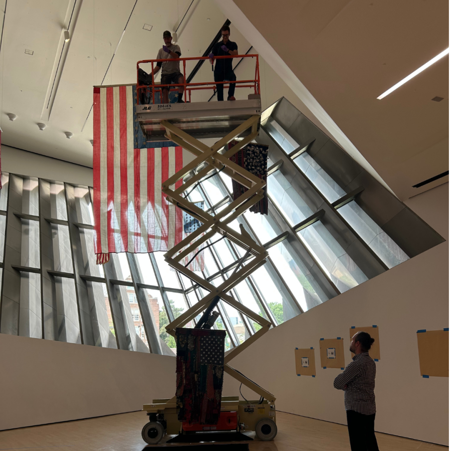 Two men on a raised lift in an art gallery decorated with American flags with a man below guiding the placement of the flag