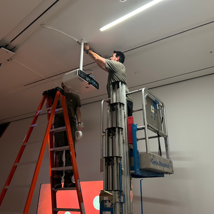 A man on a ladder works on a ceiling-mounted projector.