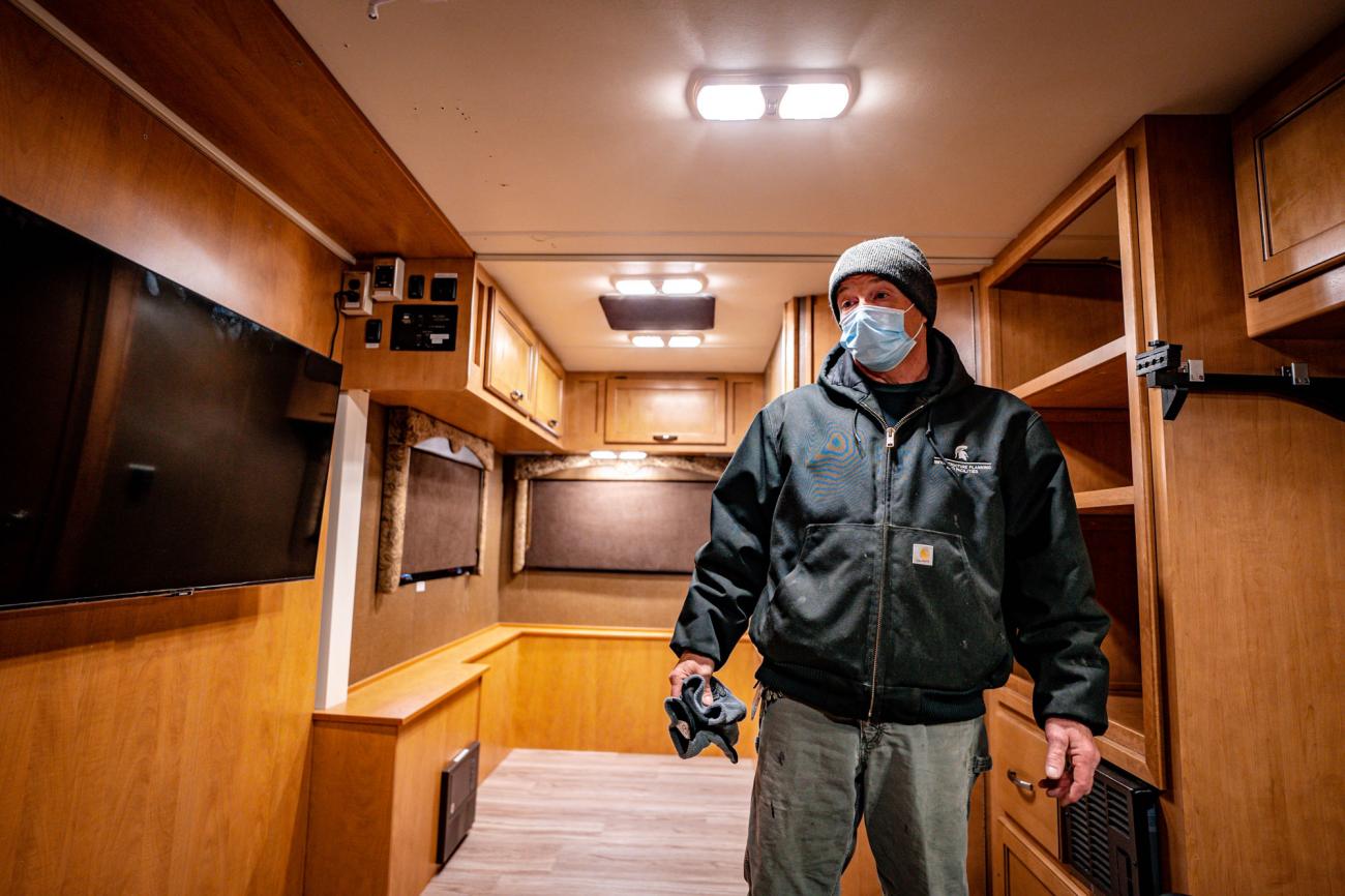 Man in blue mask and black coat stands in the light wood colored interior of the RV
