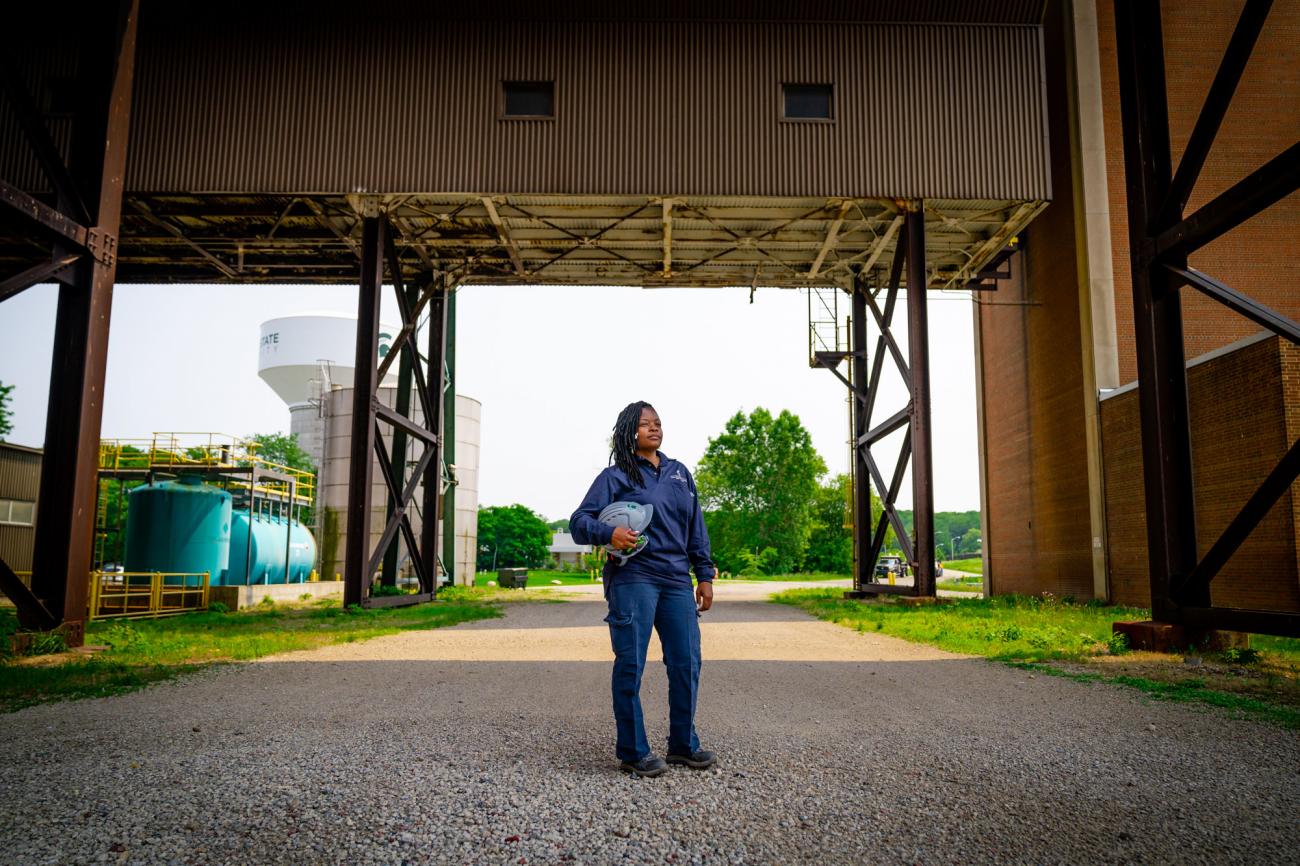 A black woman in navy blue work clothes stands outside the power plant holding a hard hat.