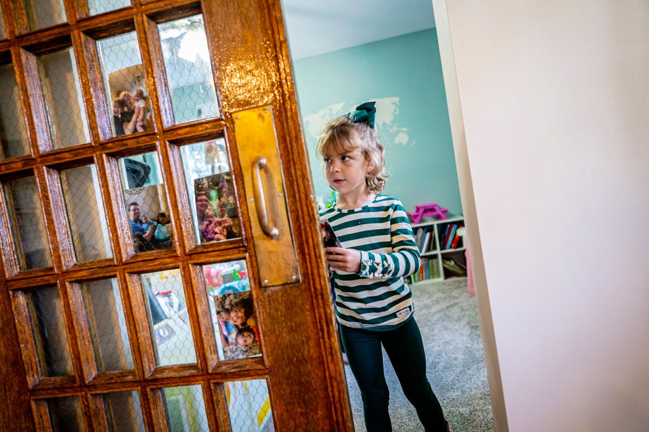 A young girl in a green and white striped shirt and green bow places family photos in the windows of a large wooden door.