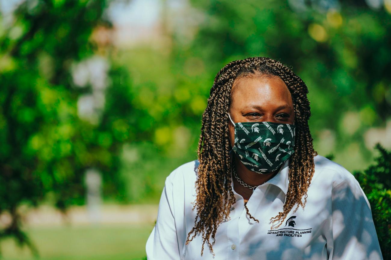 Kisha wearing a white IPF shirt and a green MSU face covering, sitting outside in dappled sunlight