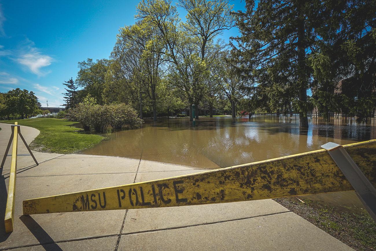 A sunny photo of MSU's campus with half the sidewalk under water from flooding