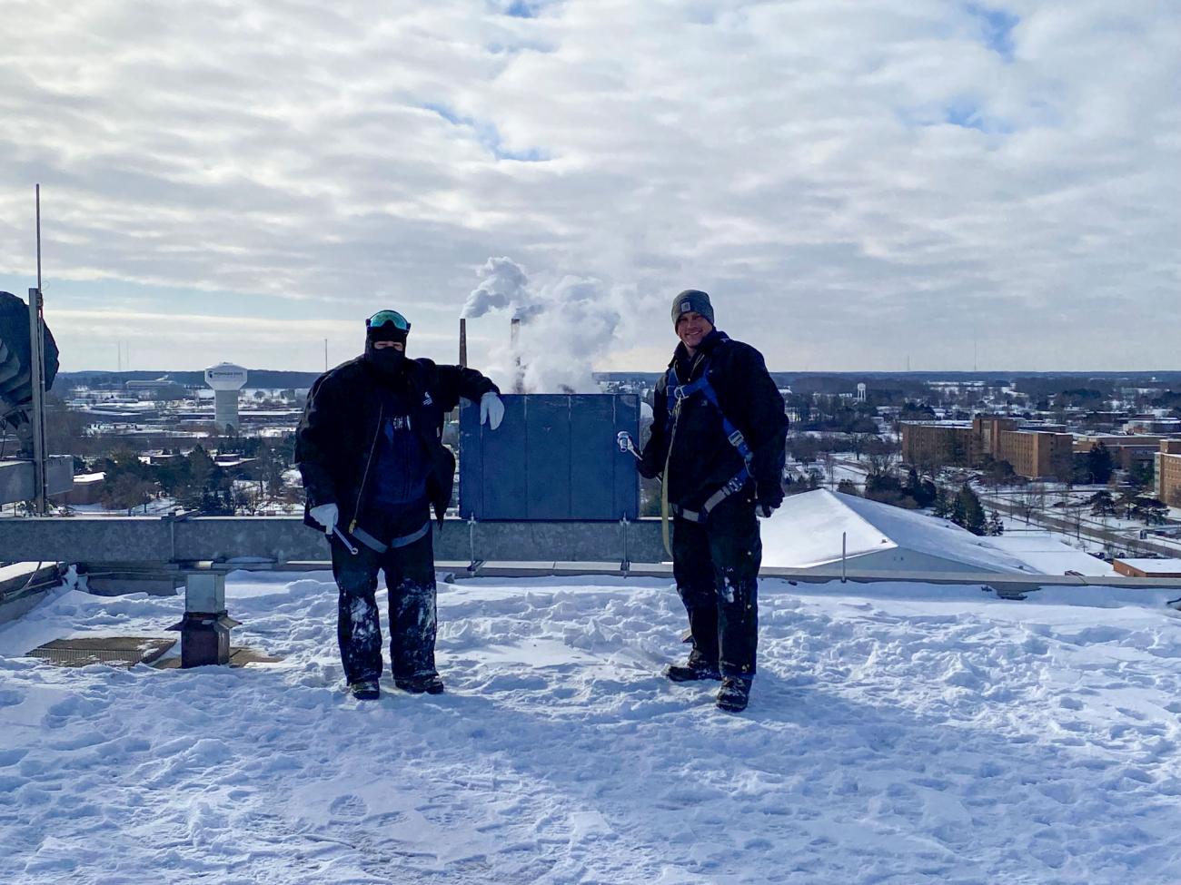 two men in cold weather gear smile on a snowy roof next to a pine box