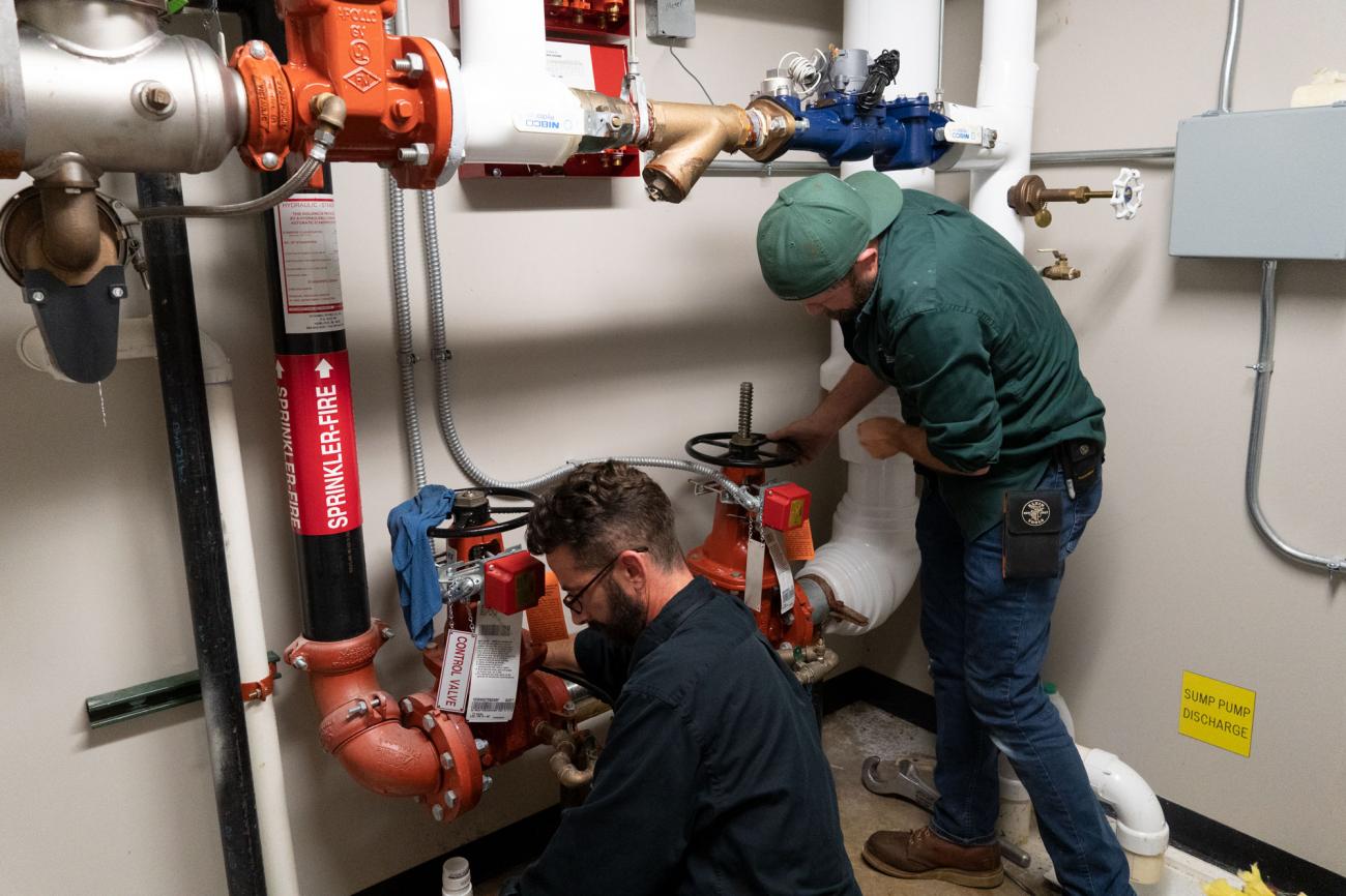 Two men working on complex plumbing installations in a mechanical room