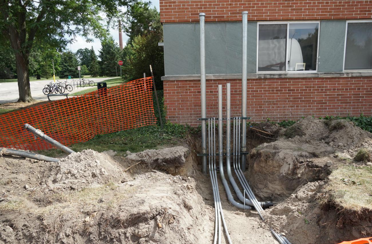 An excavated area next to a building with large metal conduits being installed