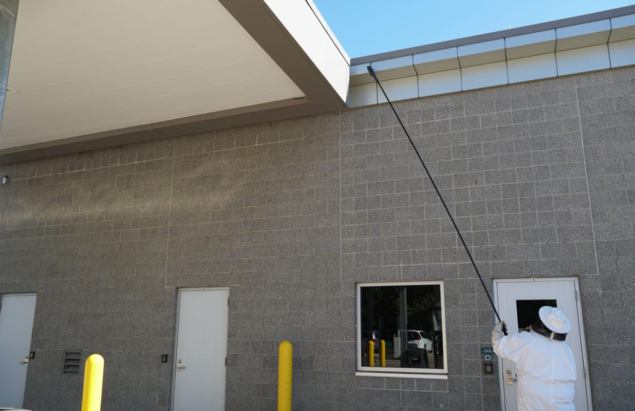 A man in a beekeeping suit lifts a large pole between a building and the roof of a fueling station