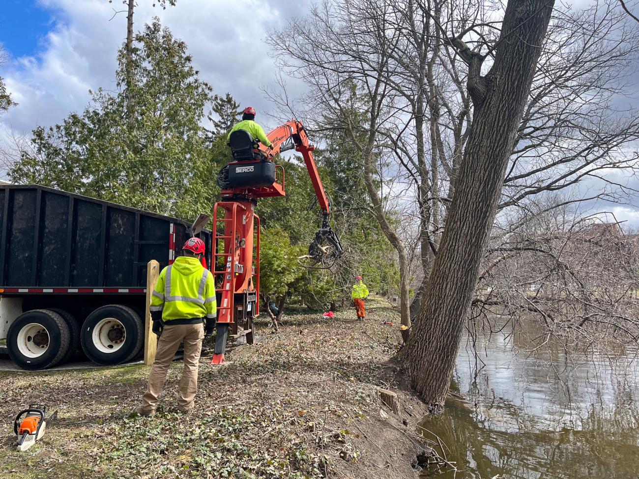 Workers use heavy equipment to remove tree debris from the bank of the Red Cedar River.