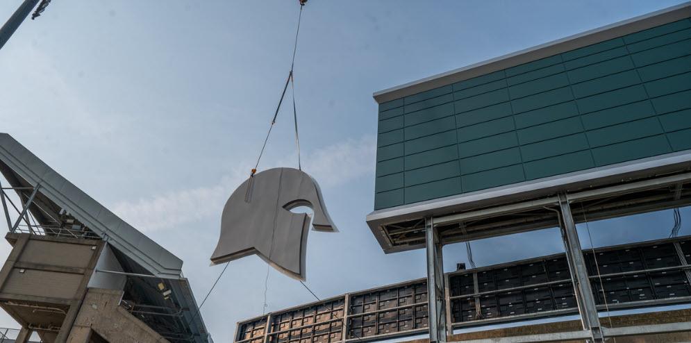 The Spartan helmet sans plume is lifted into the air by crane to be attached to the back of a new scoreboard