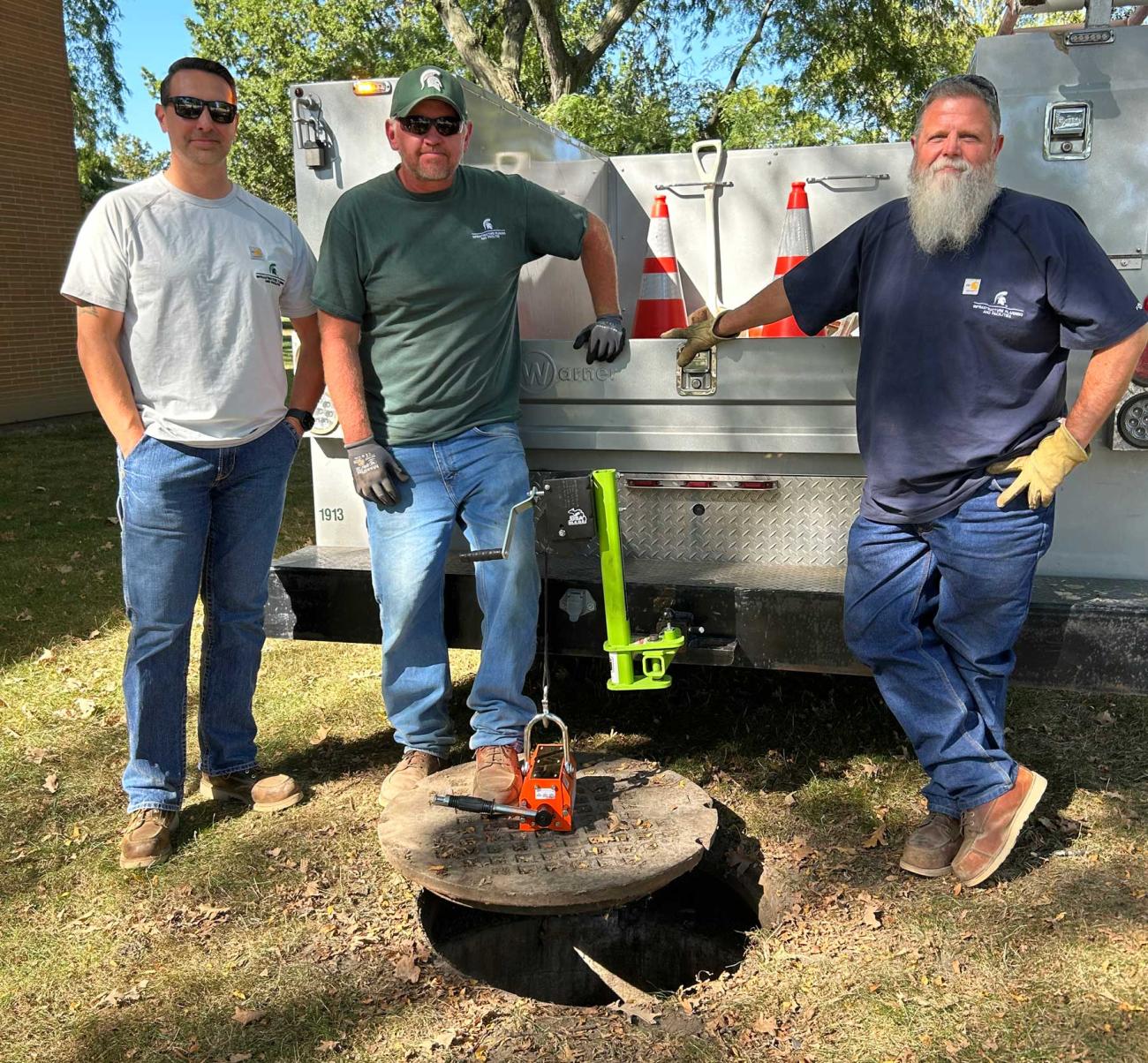 Aaron Starmer, Jim Davis, and Tom Follen pose in front of the tail of a truck that's hoisting a manhole cover