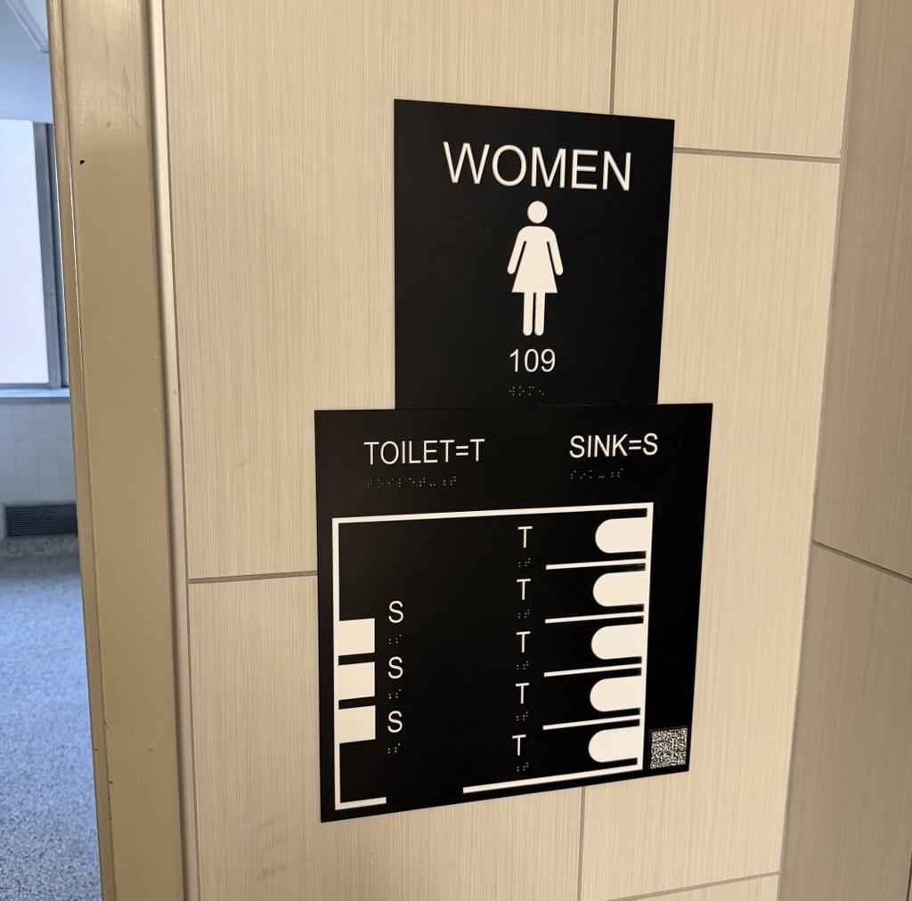 Black Accessible bathroom signage pictured on a tan wall in Bessey Hall