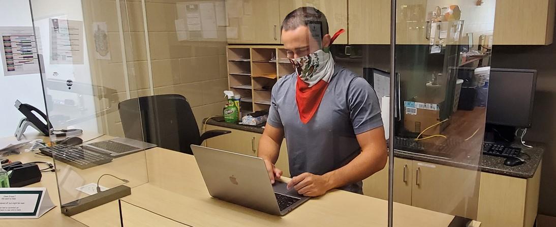 Photo of IPF IT student employee Brant Salinaz working on laptop behind plexiglass shield