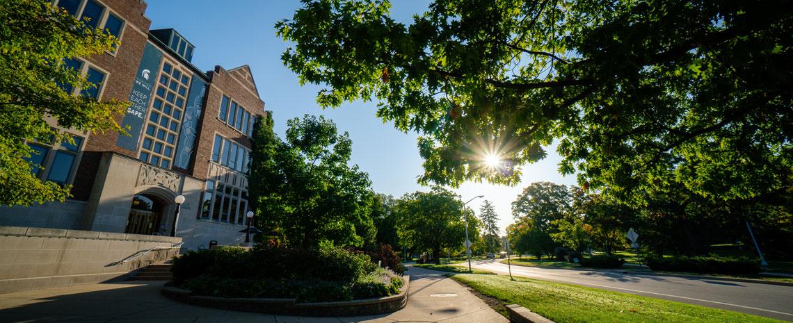 MSU's campus on a sunny day with lush green trees and foliage framing a brick building