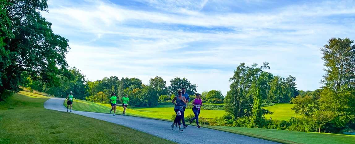 5k participants running on a paved path amongst green trees, grass and a blue sky